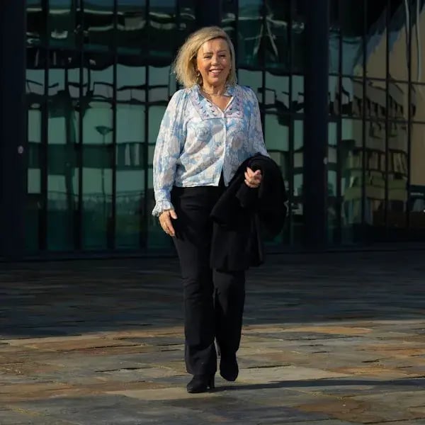 Een vrouw in een blouse en zwarte broek loopt lachend voor een glazen gebouw.