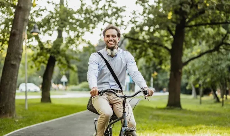 Man with dark hair in a park, riding a bicycle and smiling at the camera.
