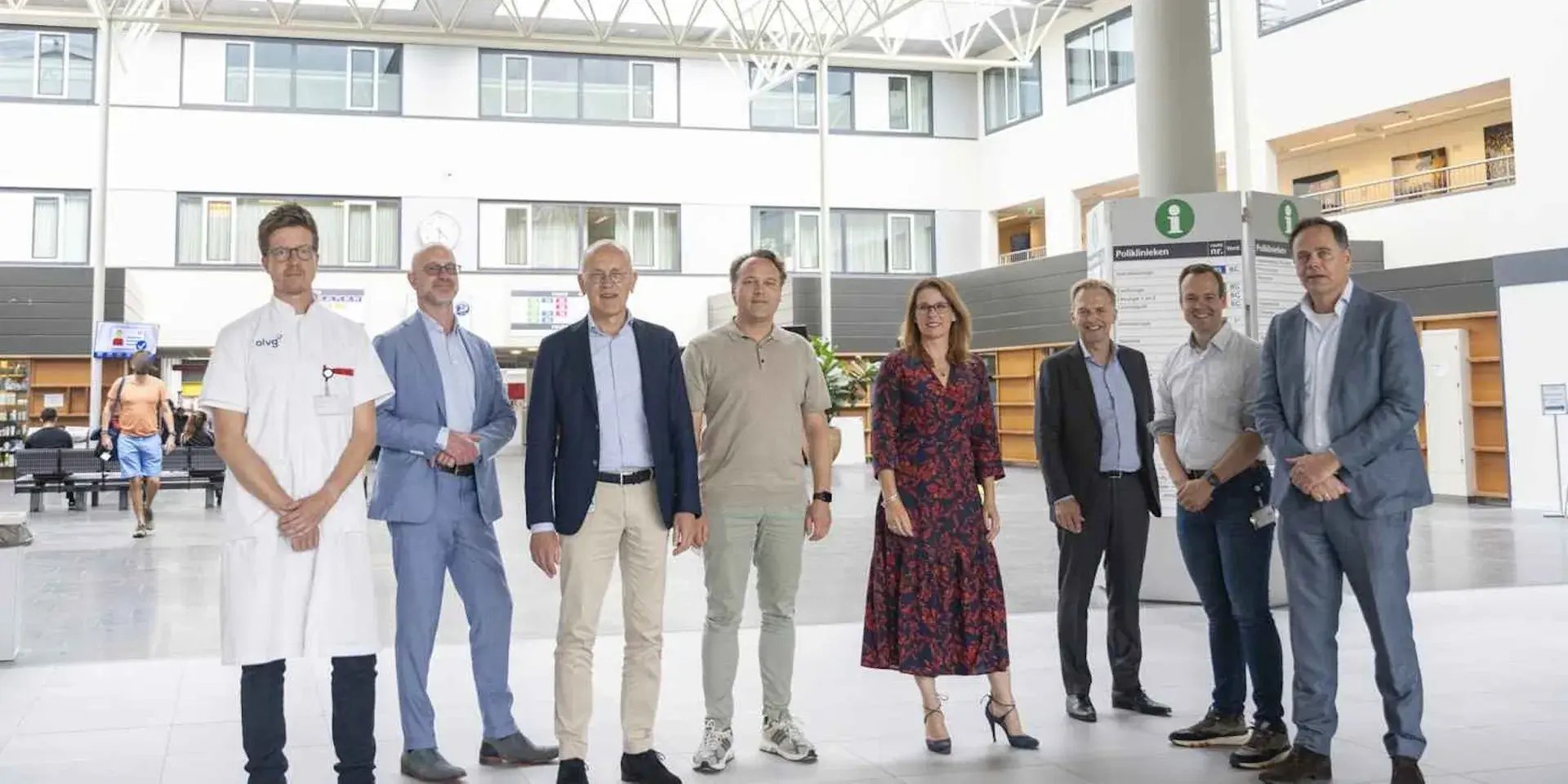 Groepsfoto van acht mensen, waaronder een arts in witte jas en anderen in zakelijke kleding, poserend in de hal van een ziekenhuis.