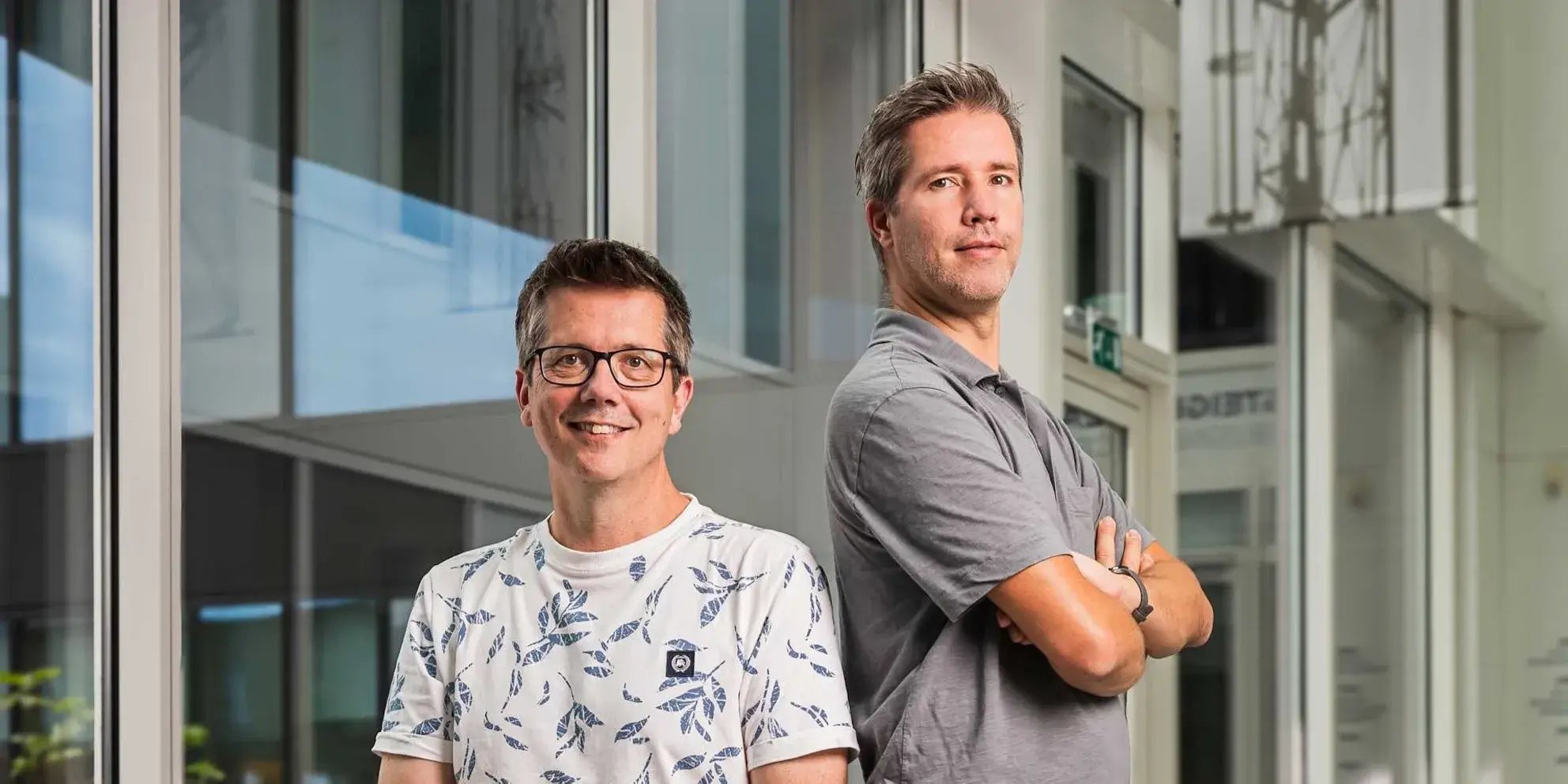 Twee collega’s staan naast elkaar, één met de armen gekruist, in een lichte kantoorruimte.