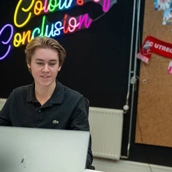 Jonge man werkt achter een laptop met een kleurrijk neonbord op de achtergrond.