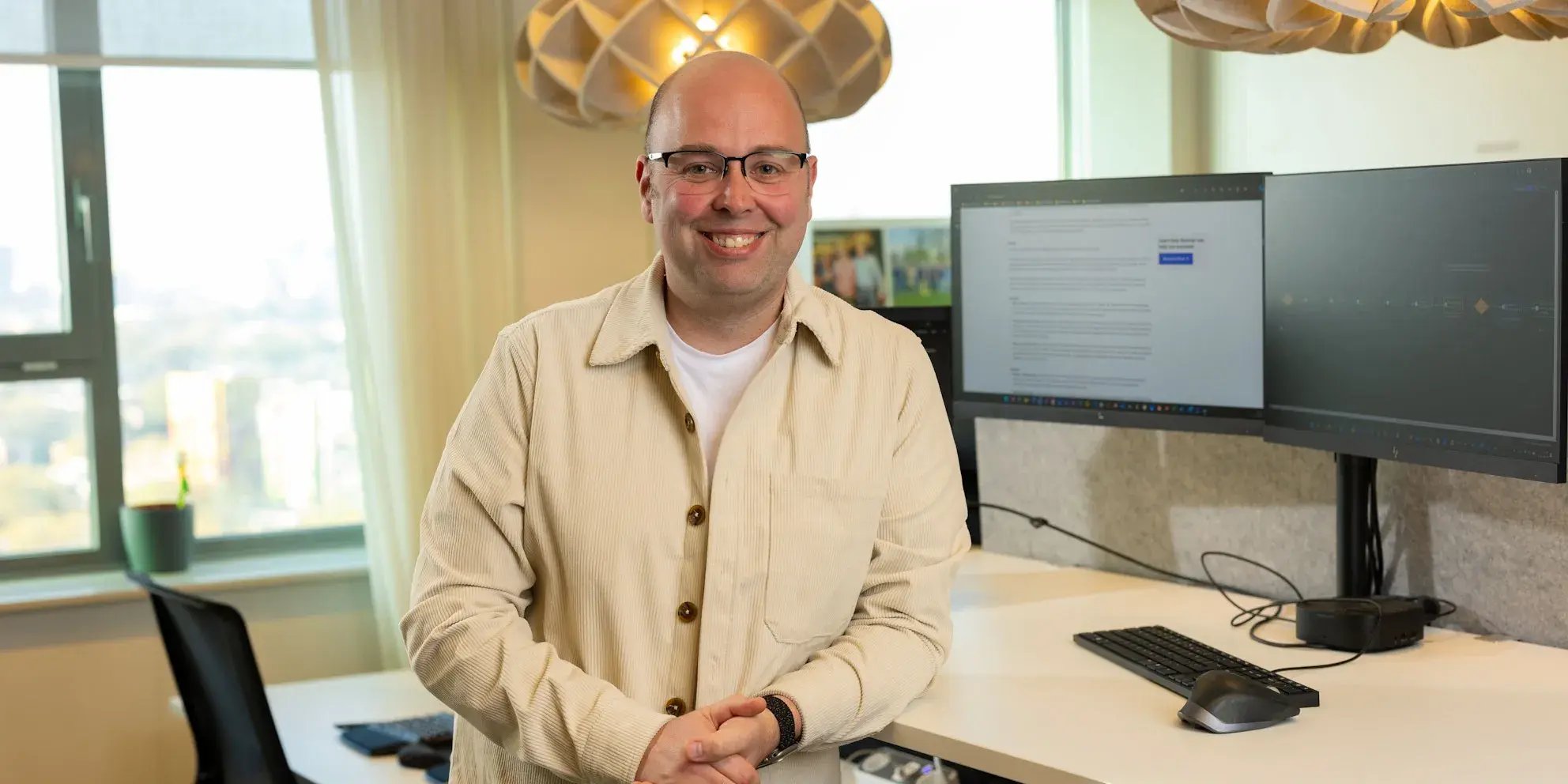 Glimlachende man met bril in een beige overhemd staat in een kantoor naast twee computerschermen.