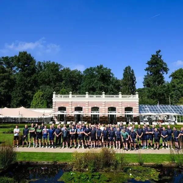 Een grote groep wielrenners poseert voor een statig gebouw in een groene tuin op een zonnige dag.