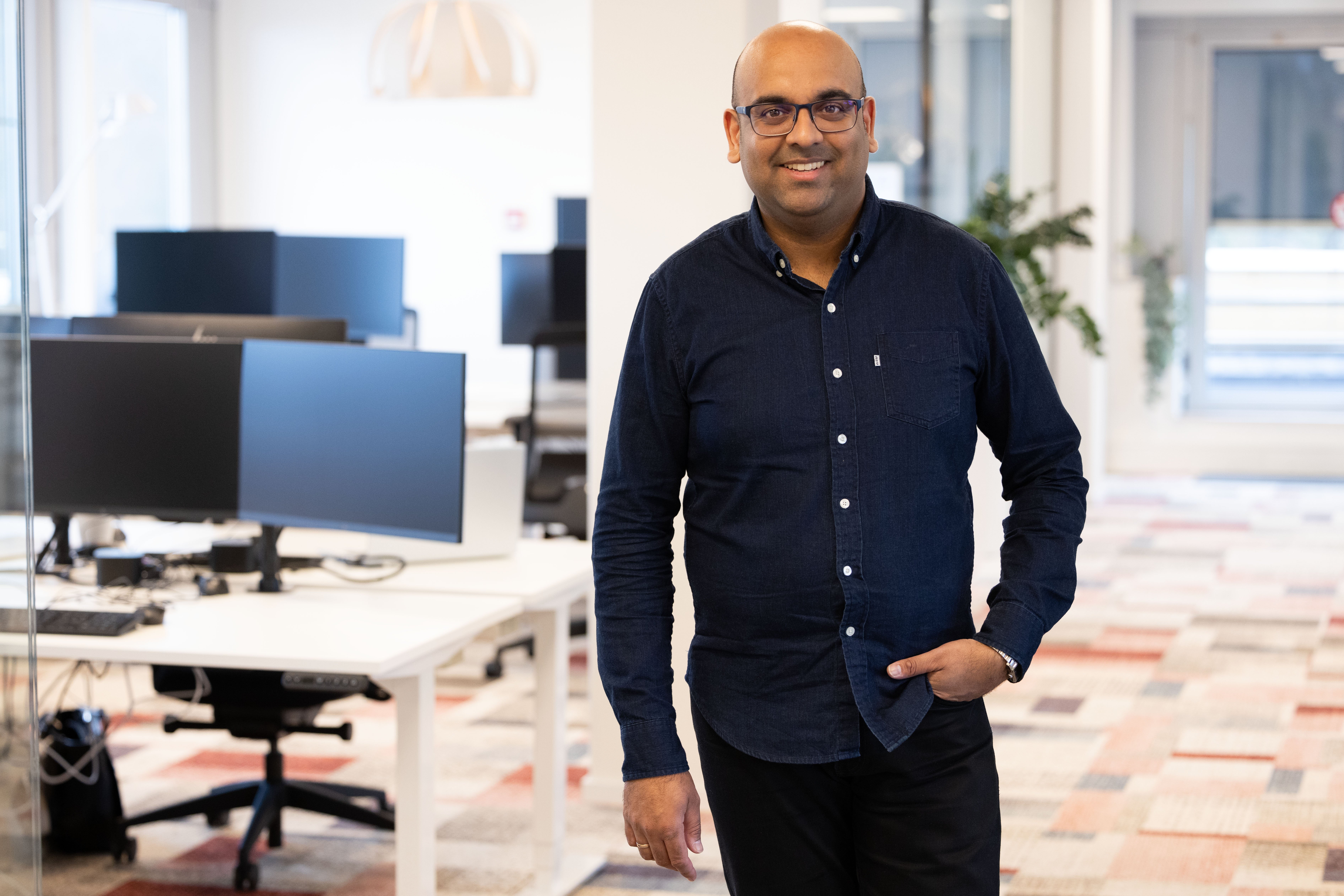 Man in een donkerblauw overhemd staat in een modern kantoor met bureaus en computers.