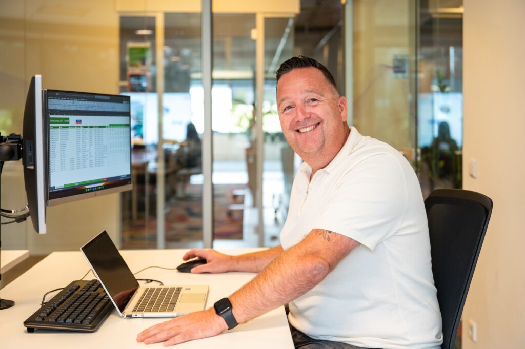 Een collega zit aan zijn bureau en lacht vriendelijk.