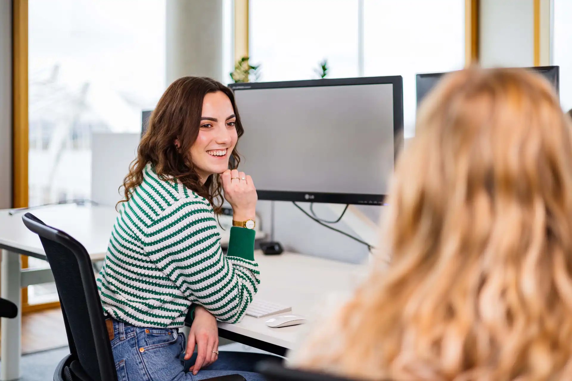 Two female colleagues smiling while talking at a desk with a computer.