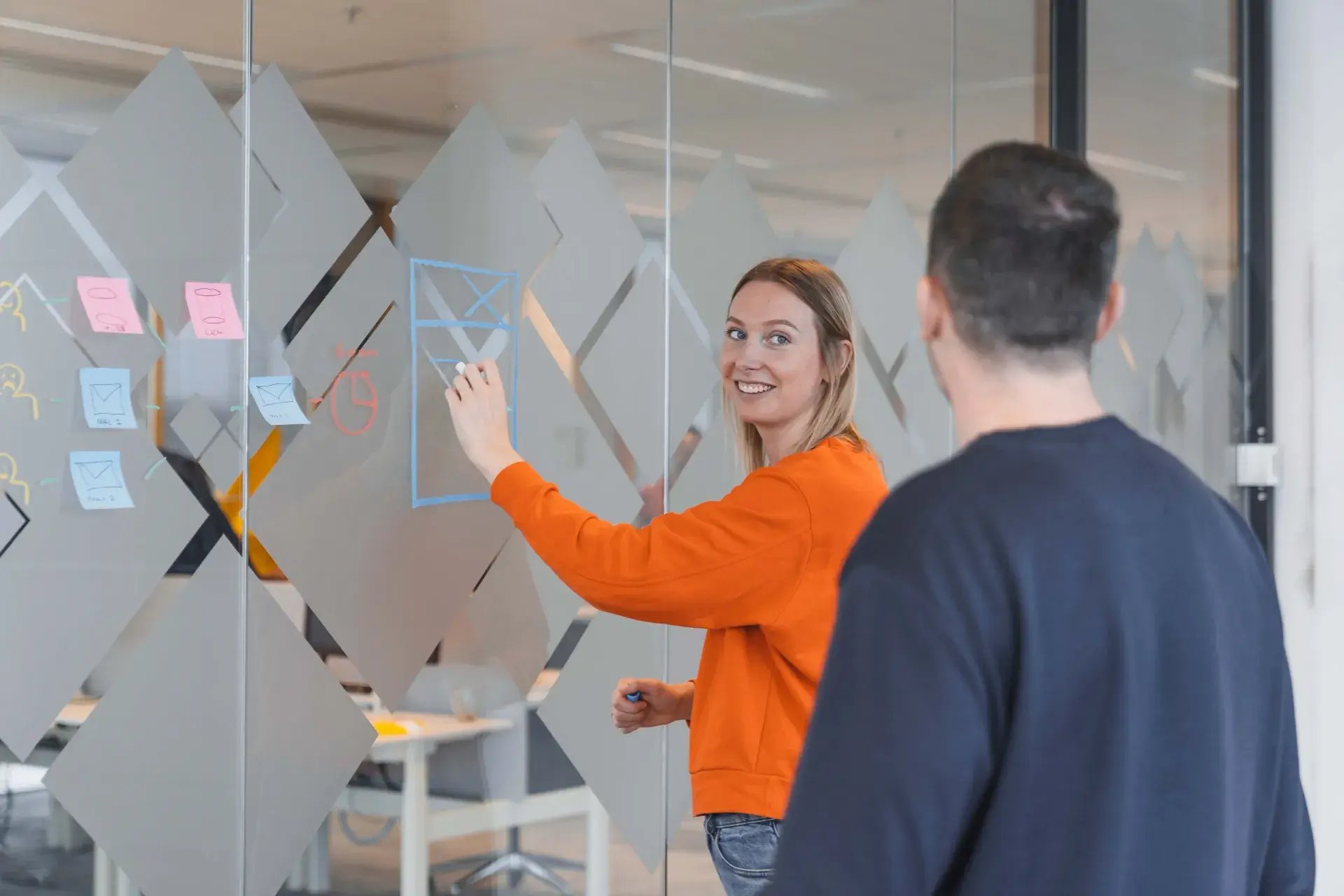 Een vrouwelijke collega tekent een model op een glazen wand en laat het zien aan een mannelijke collega.