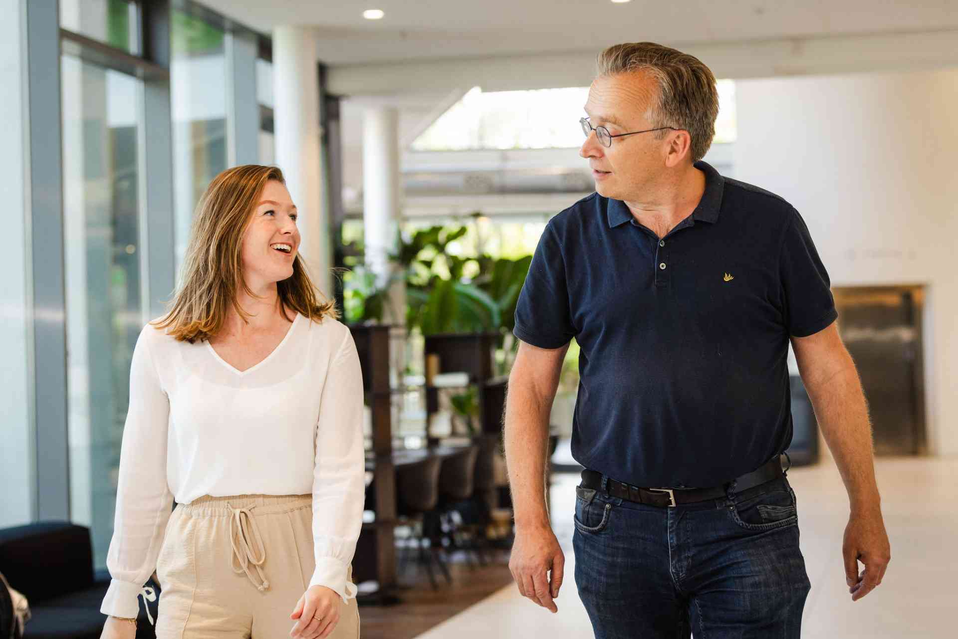Twee collega's lopen door het kantoor en lachen naar elkaar.