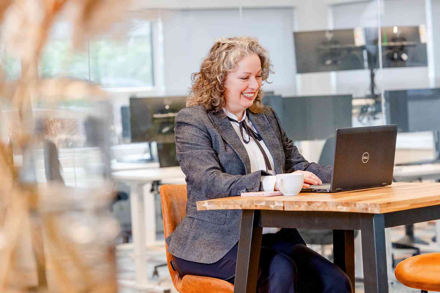 Een vrouw zit aan een hoge tafel met haar laptop en lacht vrolijk.