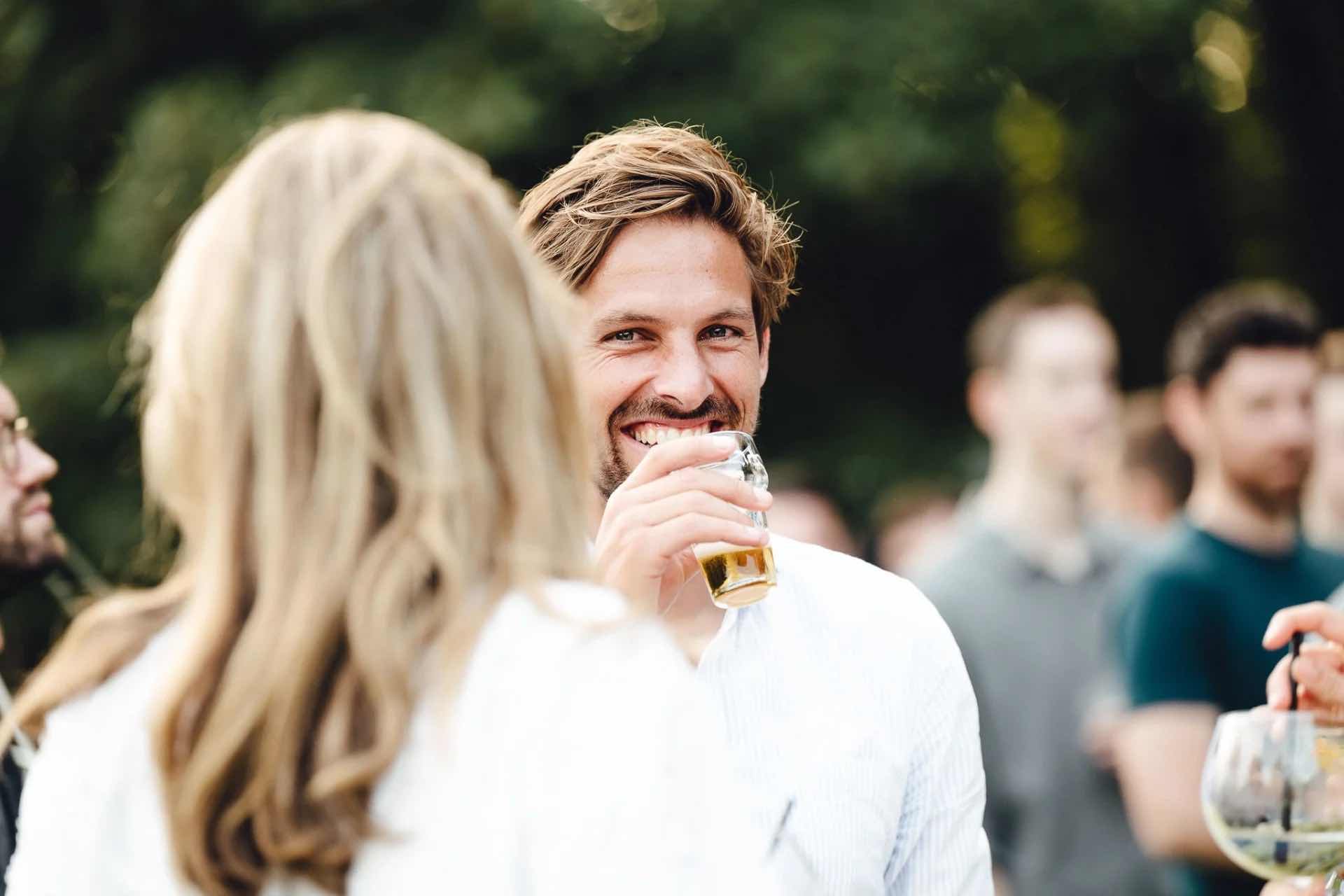 Een man met een glimlach houdt een glas bier vast terwijl hij met iemand in gesprek is op een evenement.