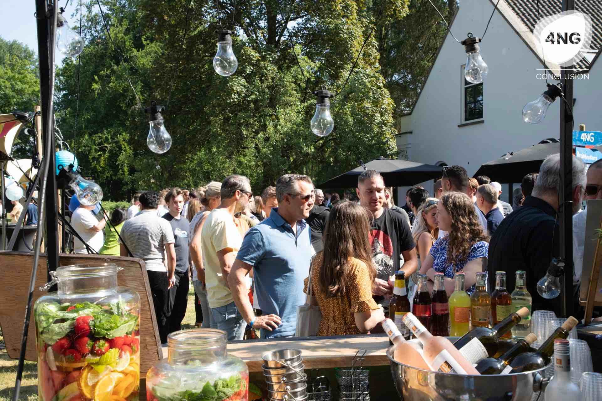 Mensen staan in een tuin bij een bar, met kleurrijke verlichting en een frisse zomerse sfeer.