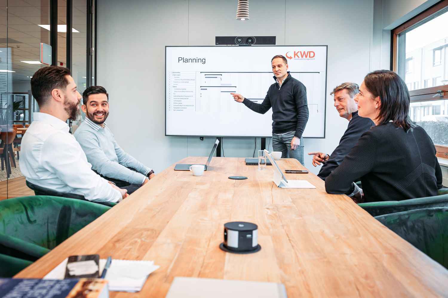 Collega's zitten aan tafel in een vergaderruimte en luisteren naar een collega die wijst naar het scherm.