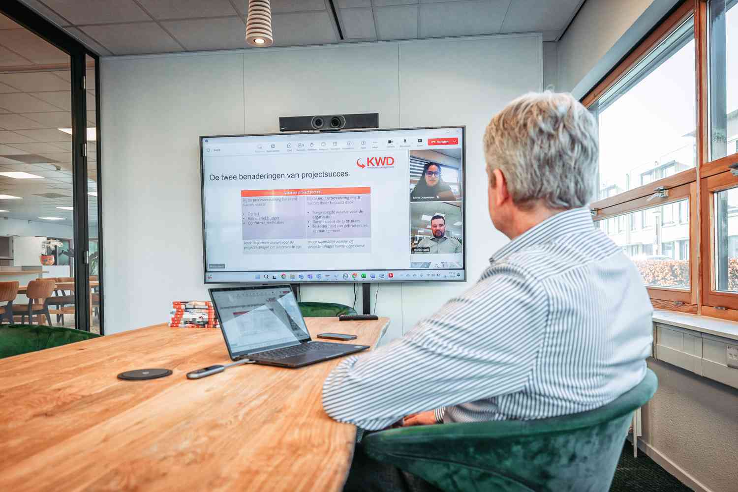 Een man zit aan een lang bureau en kijkt naar een meeting op het scherm.