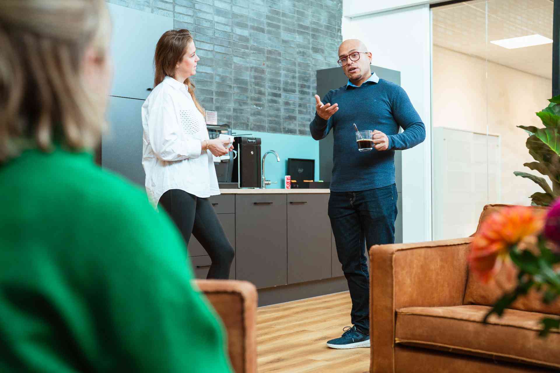 Twee mensen praten in een kantoor, terwijl een man naar een vrouw kijkt.