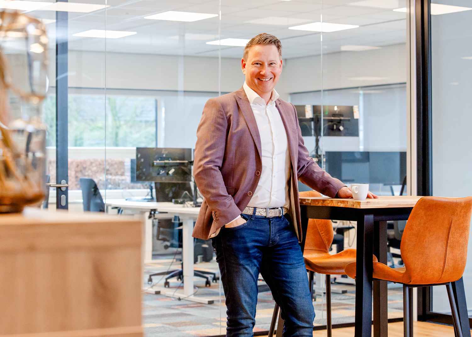 Een man leunt tegen een hoge tafel aan en kijkt lachend voor zich uit.