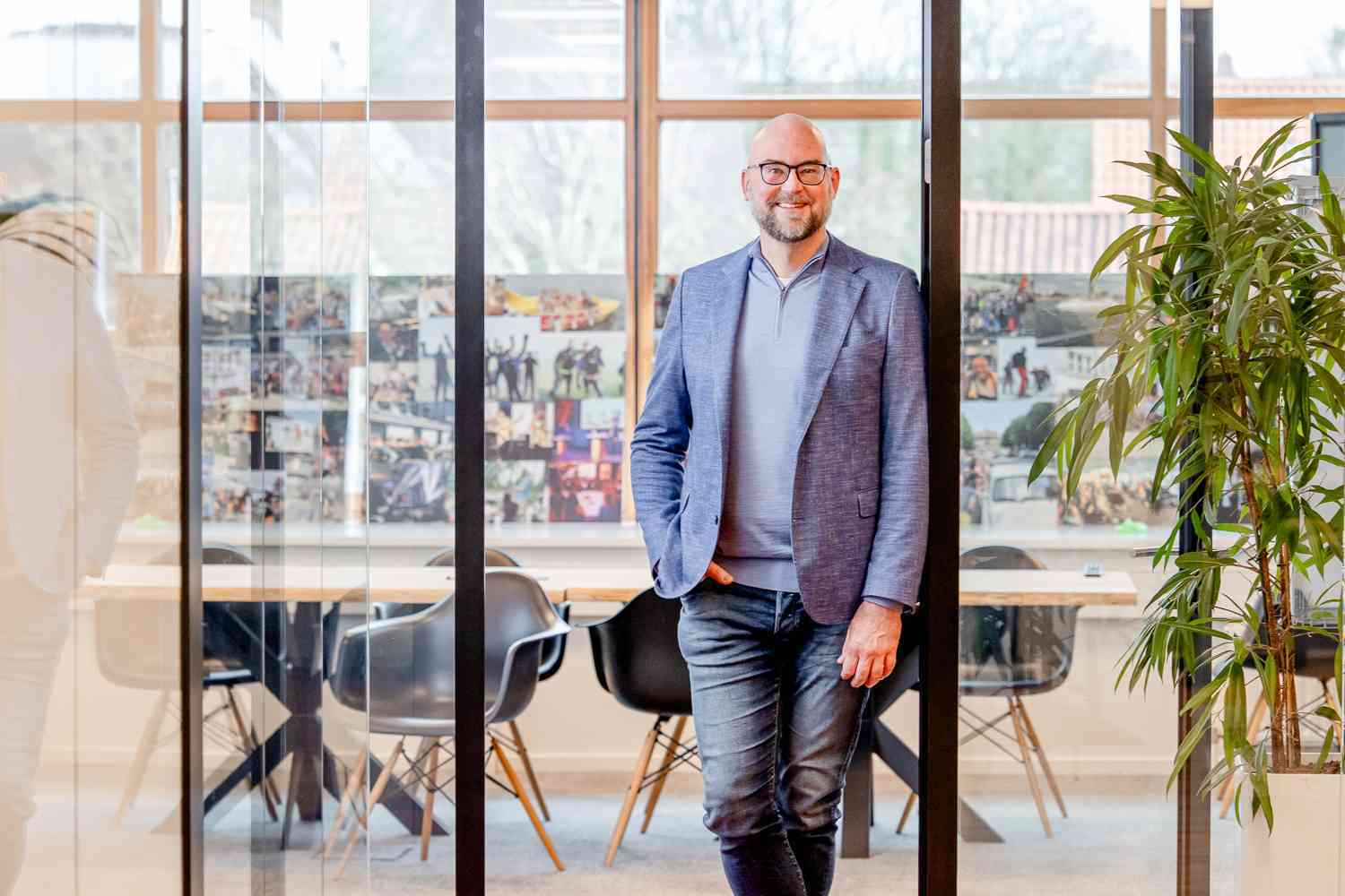 Een man leunt tegen de glazen wand van een vergaderruimte en kijkt lachend voor zich uit.