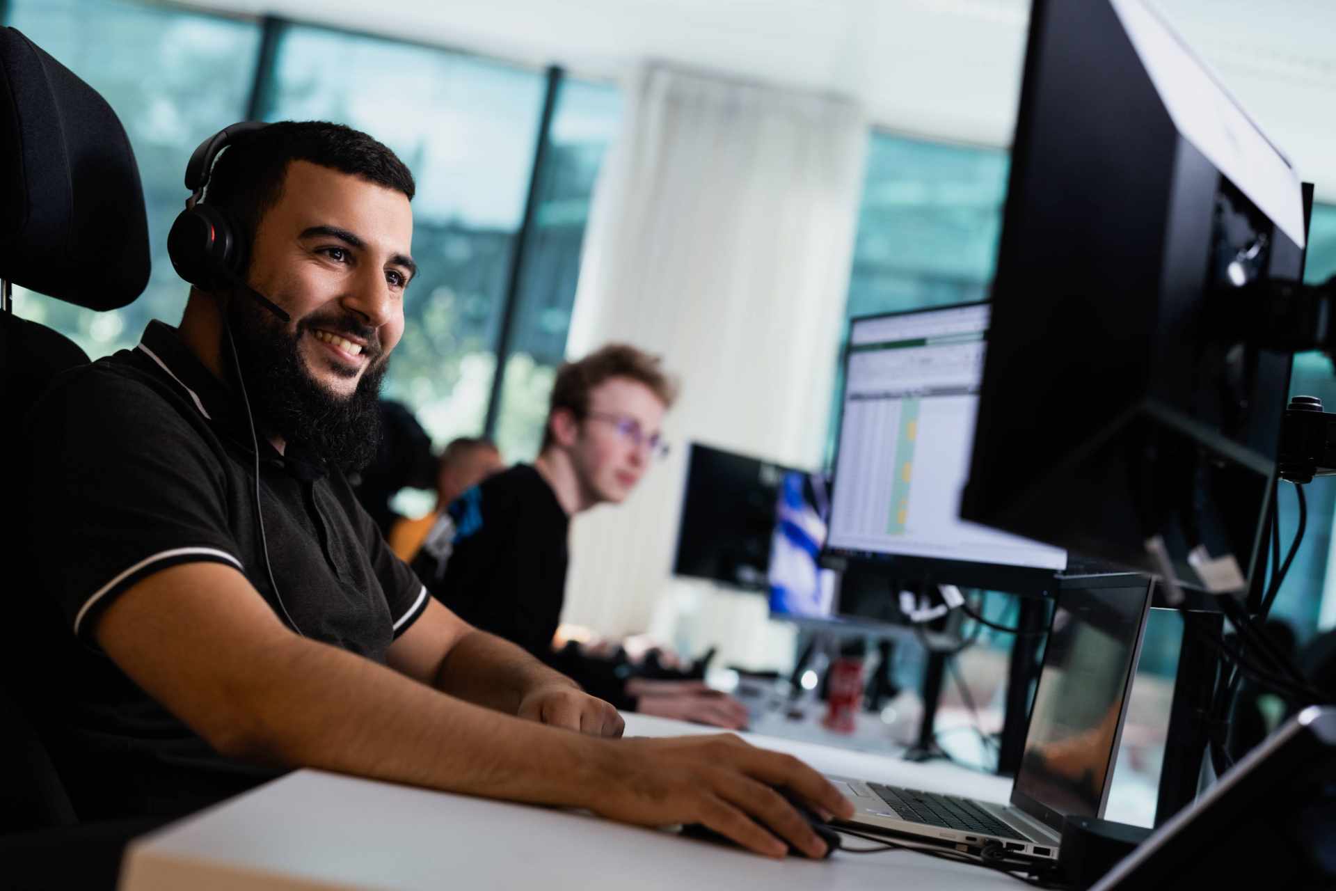 Een werknemer met een headset werkt aan een computer in een open kantoorruimte.