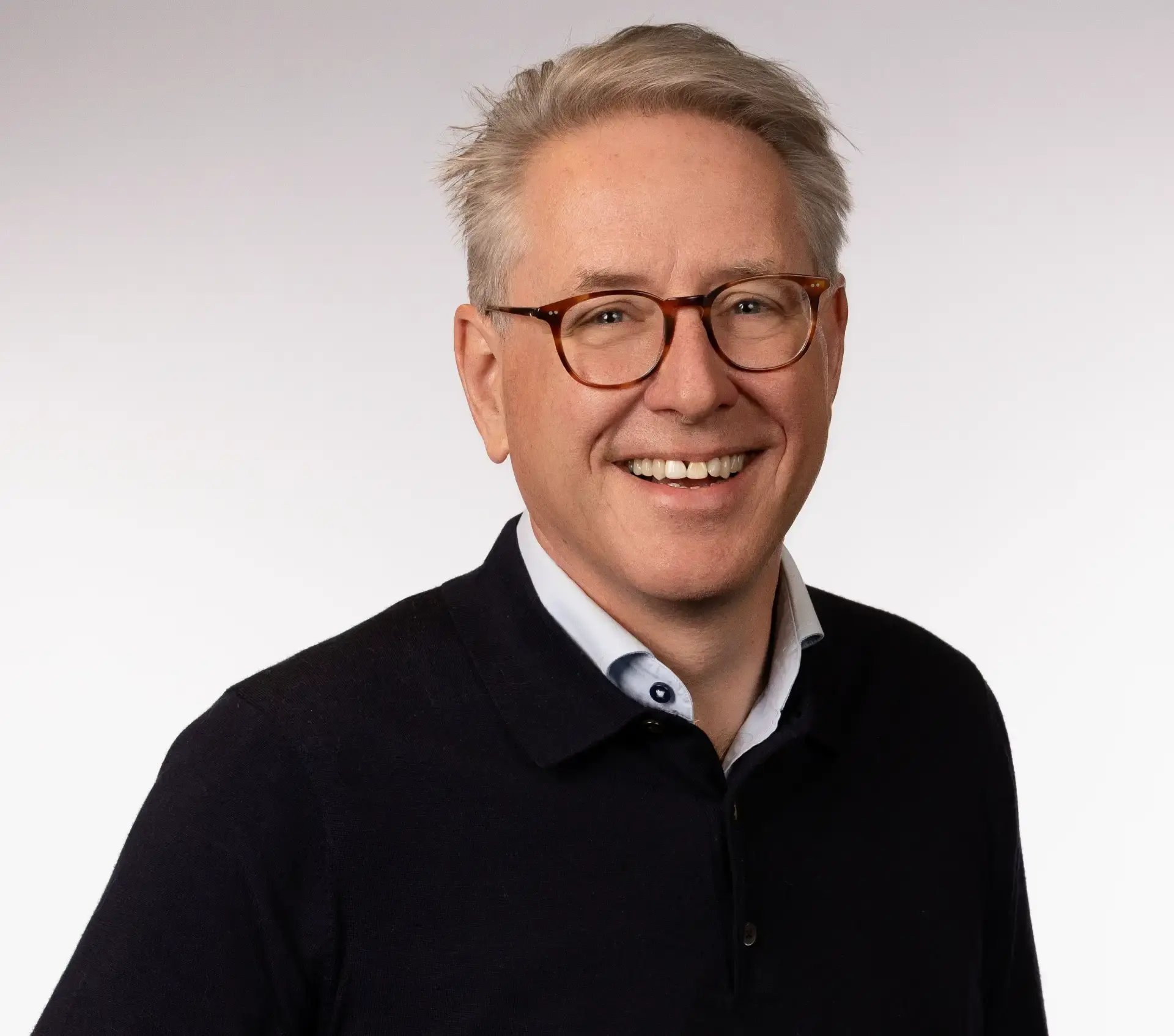 Lachende man met bril en zwarte trui kijkt in camera.