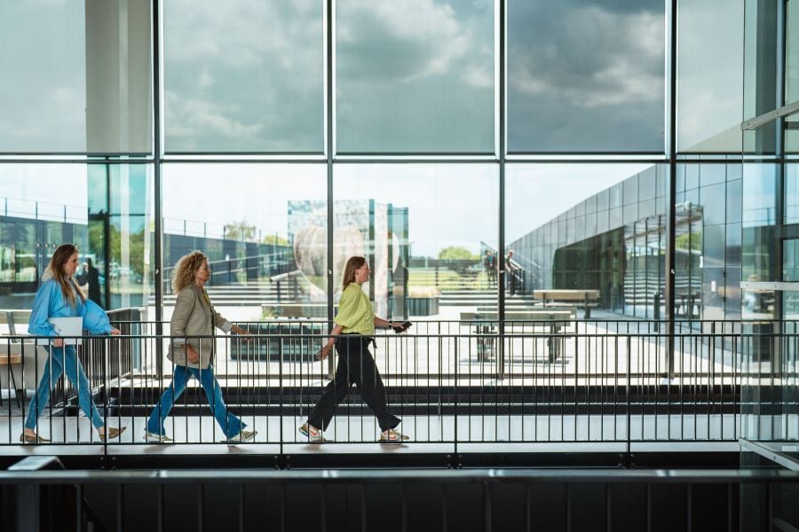 Three women walk through a modern office hallway with large windows, overlooking the outside space.