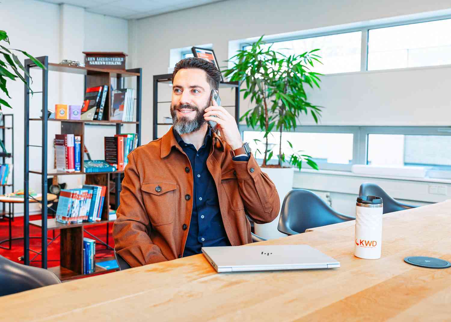 Een man zit aan een bureau in een kantoor en praat via de telefoon, met een laptop en een koffiemok voor zich.