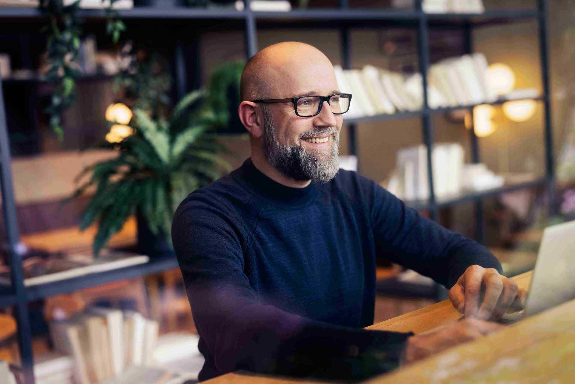 Man with glasses smiling while working on a laptop.