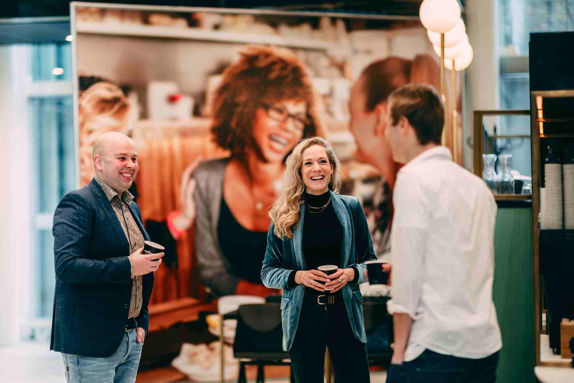 Drie mensen lachen en praten met koffie in de hand.