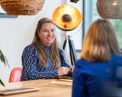 Een vrouw in een blauwe blouse lacht terwijl ze met een andere vrouw aan een tafel zit.