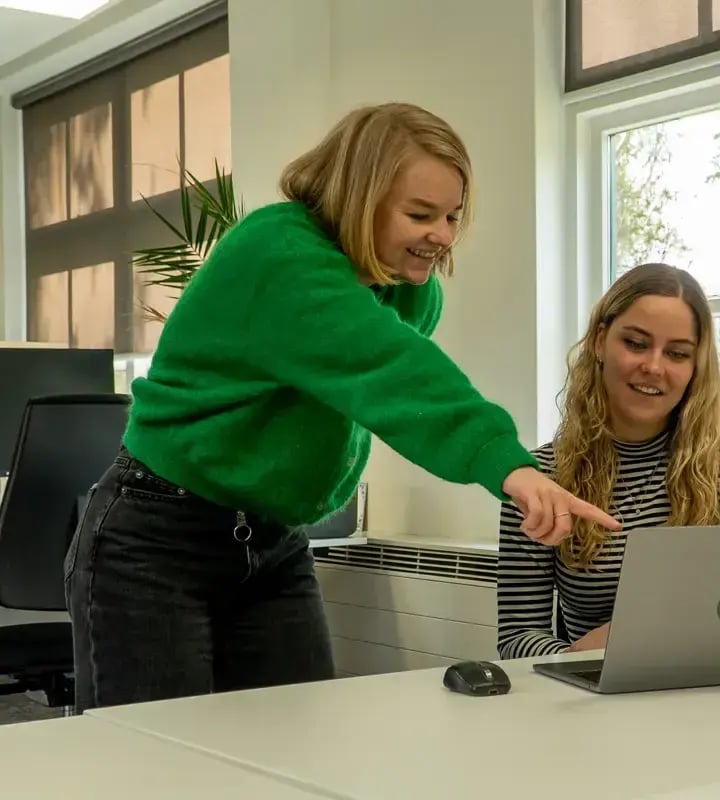 Vrouw met groene trui staat aan bureau naast andere vrouw en wijst naar laptop.