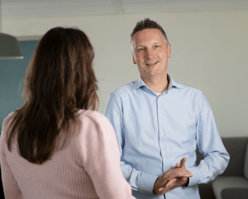 Man kijkt in camera, in gesprek met vrouw.