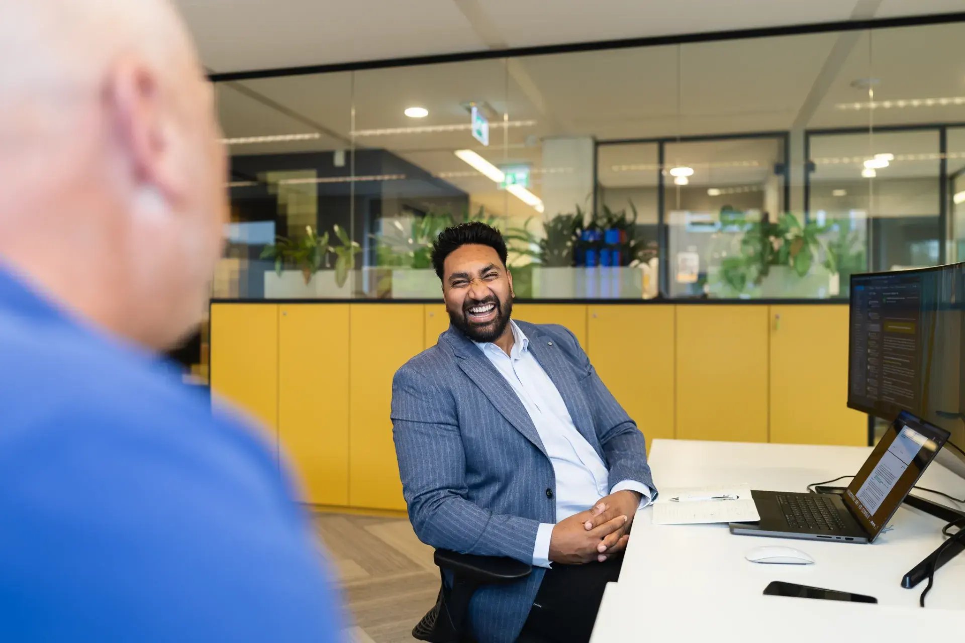Black man with a beard wearing a blazer, sitting behind a laptop, talking to a colleague.