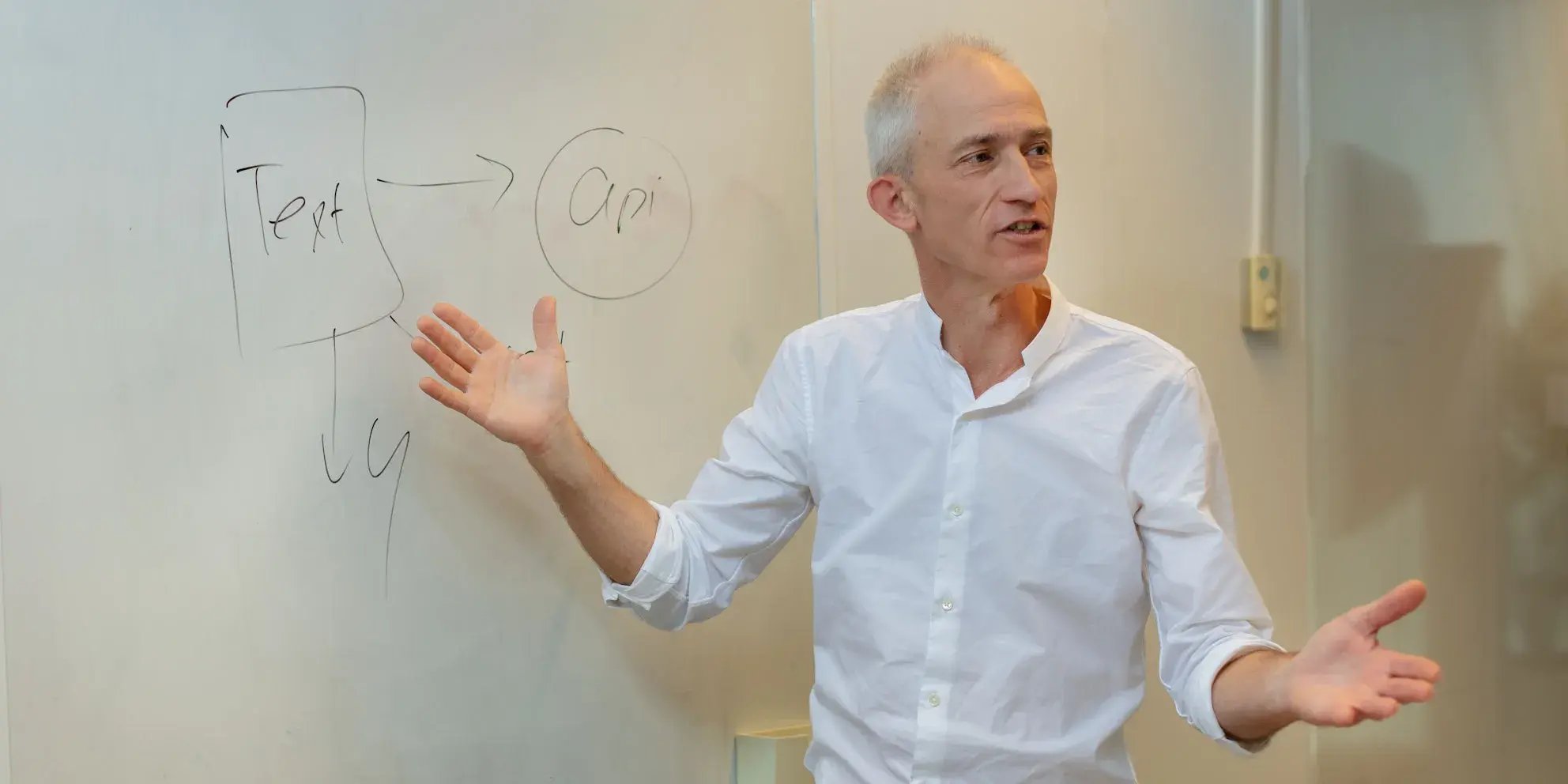 Man in witte blouse legt iets uit bij een whiteboard met de tekst 
