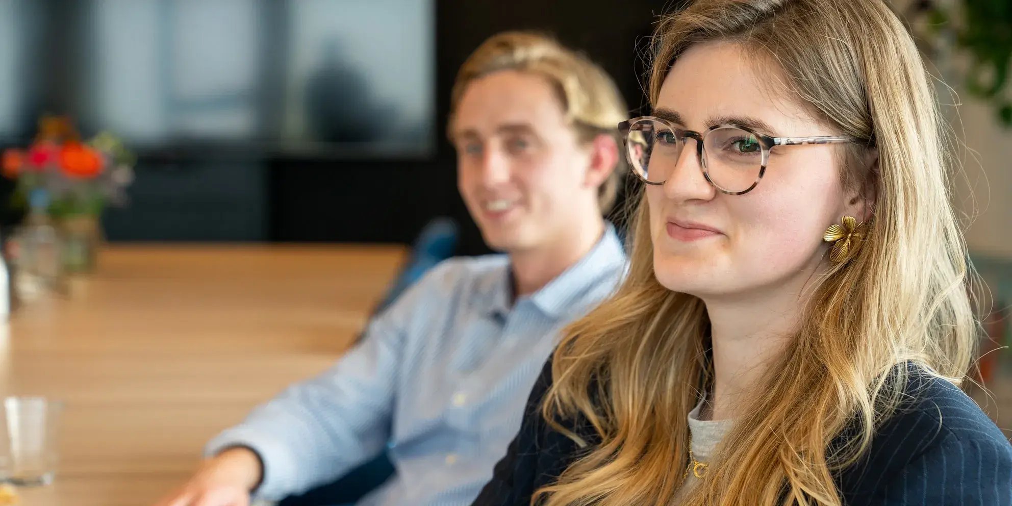 Vrouw met bril en gouden oorbellen glimlacht tijdens een vergadering, met een man op de achtergrond.