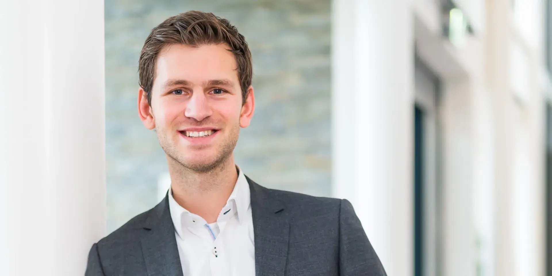 Man in pak lacht naar de camera in een lichte kantooromgeving.