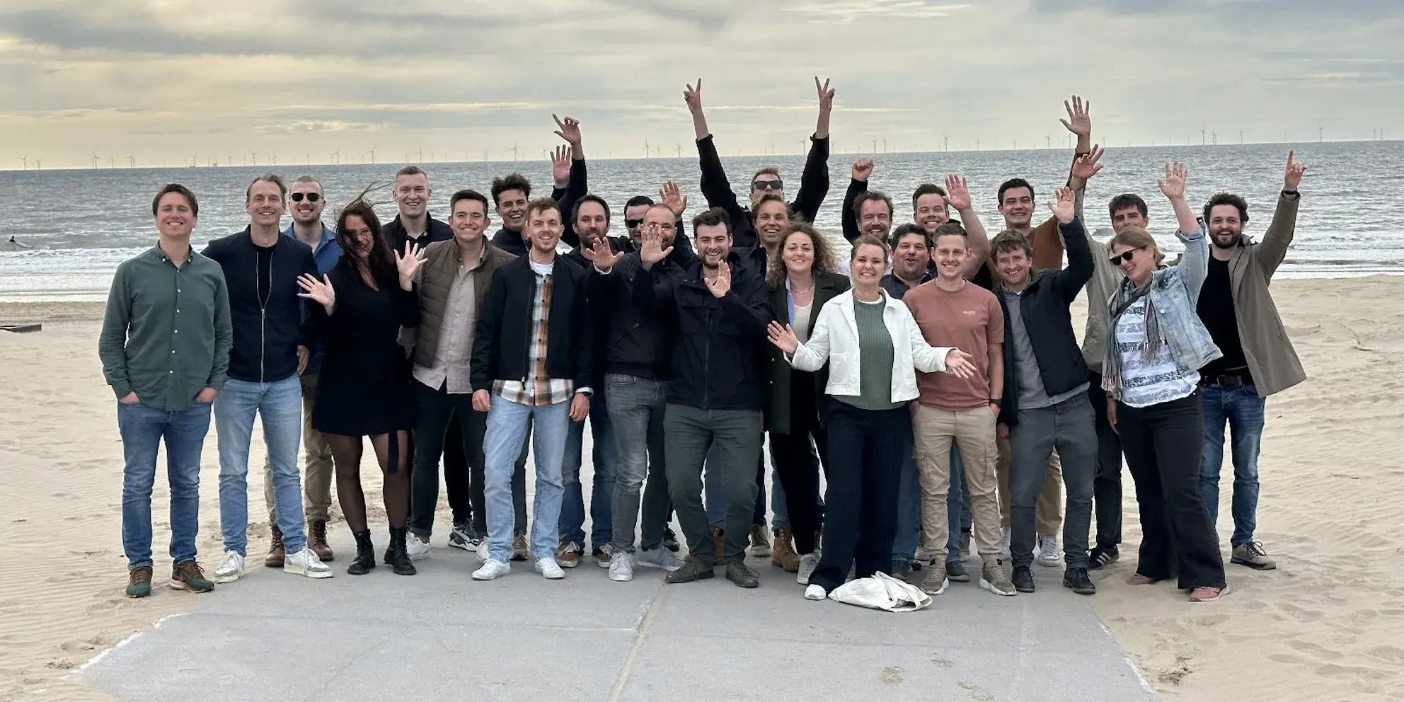 Groep mensen poseert lachend en zwaaiend op het strand bij de zee.