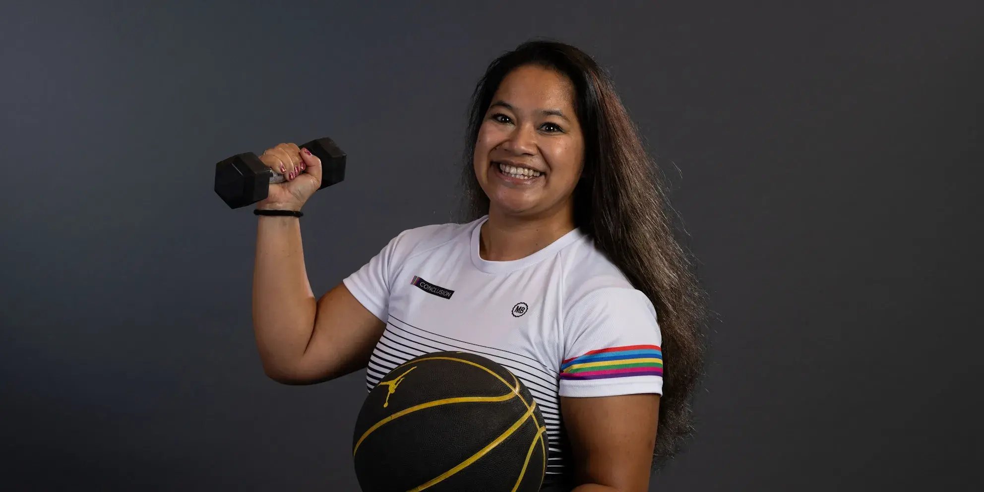 Vrouw in sportshirt glimlacht terwijl ze een dumbbell en basketbal vasthoudt.