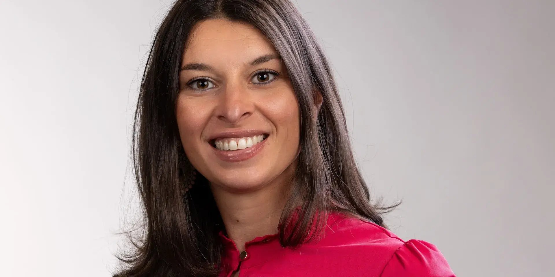 Vrouw met lang donker haar lacht in de camera en draagt een felroze blouse.