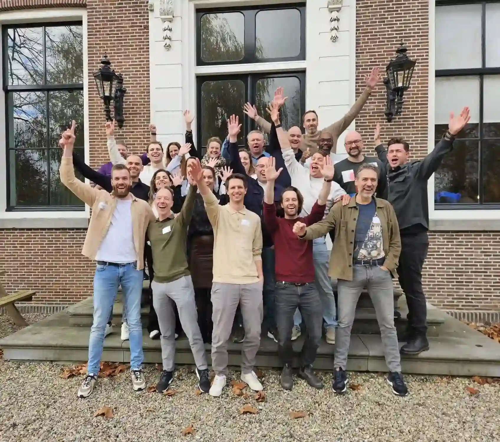 Groepsfoto van blije mensen met handen in de lucht voor een pand.