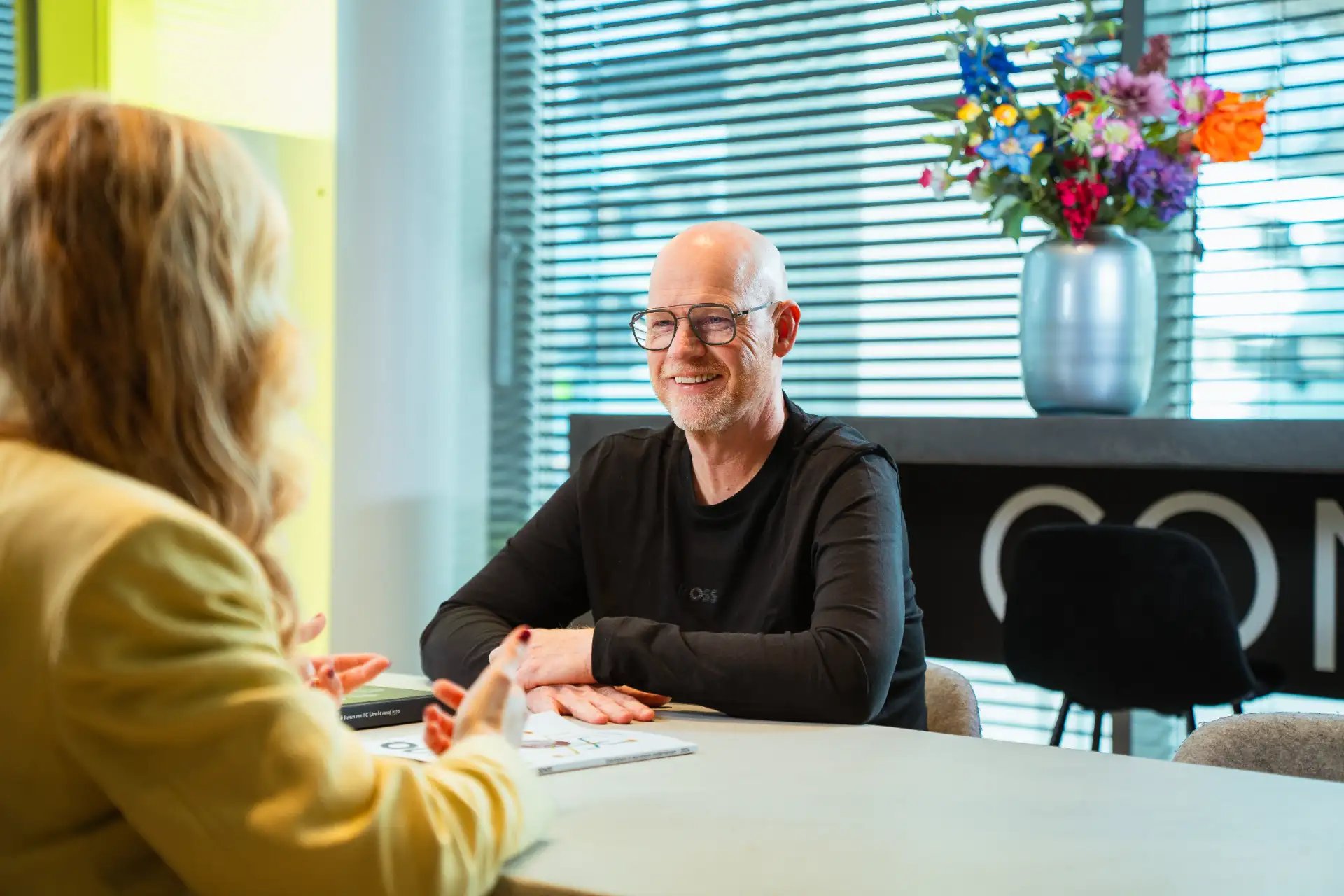 Kale man met bril in gesprek met vrouw aan tafel.