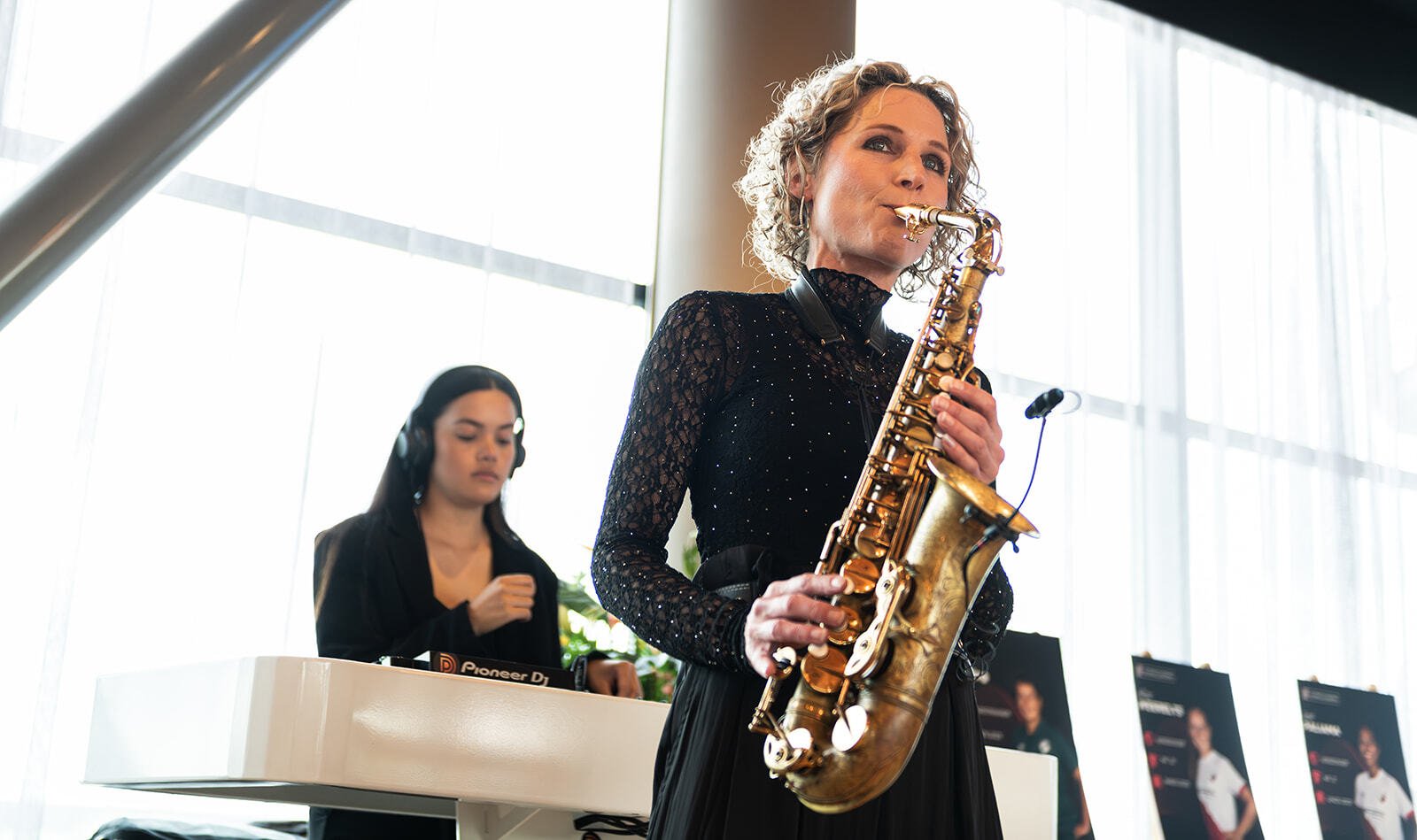 Vrouw speelt saxofoon en achter haar staat een DJ.
