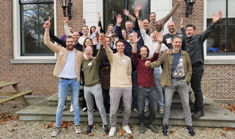 Groepsfoto van blije mensen met handen in de lucht voor een pand.