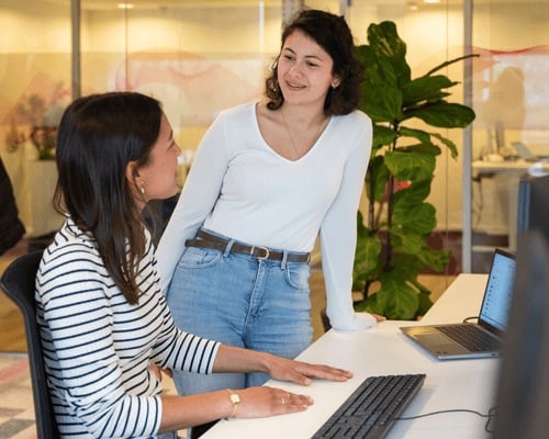 Een vrouw staat aan het bureau van haar collega en zegt iets.