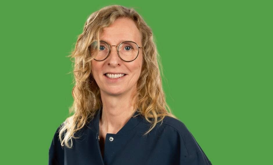 A woman with curly blonde hair and glasses smiles in front of a green background.