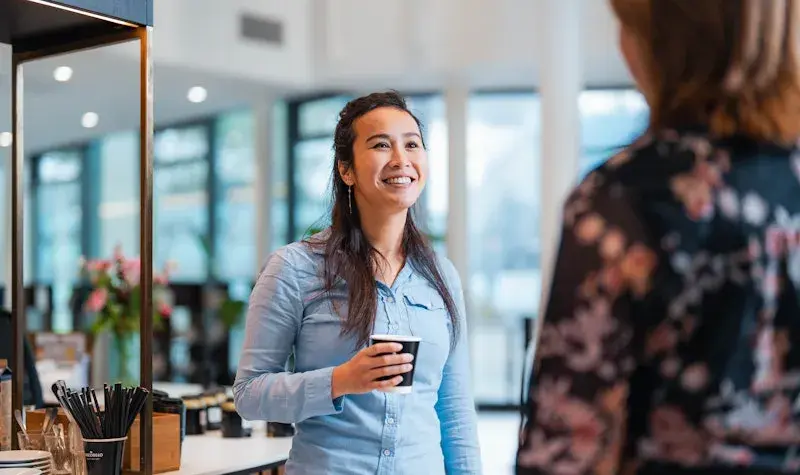 Vrouw met donker haar lachend naar collega met koffie in haar hand