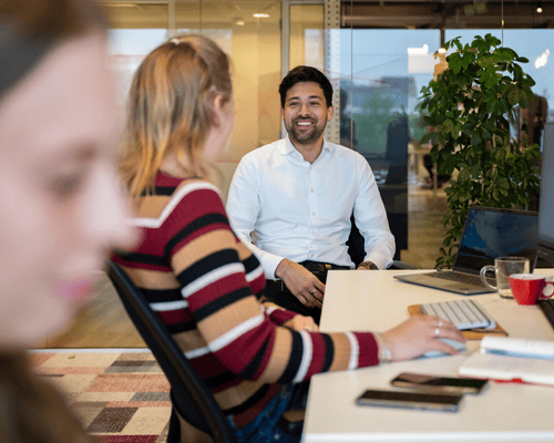 Een man lacht terwijl hij aan een bureau zit met een vrouw in gestreepte trui.